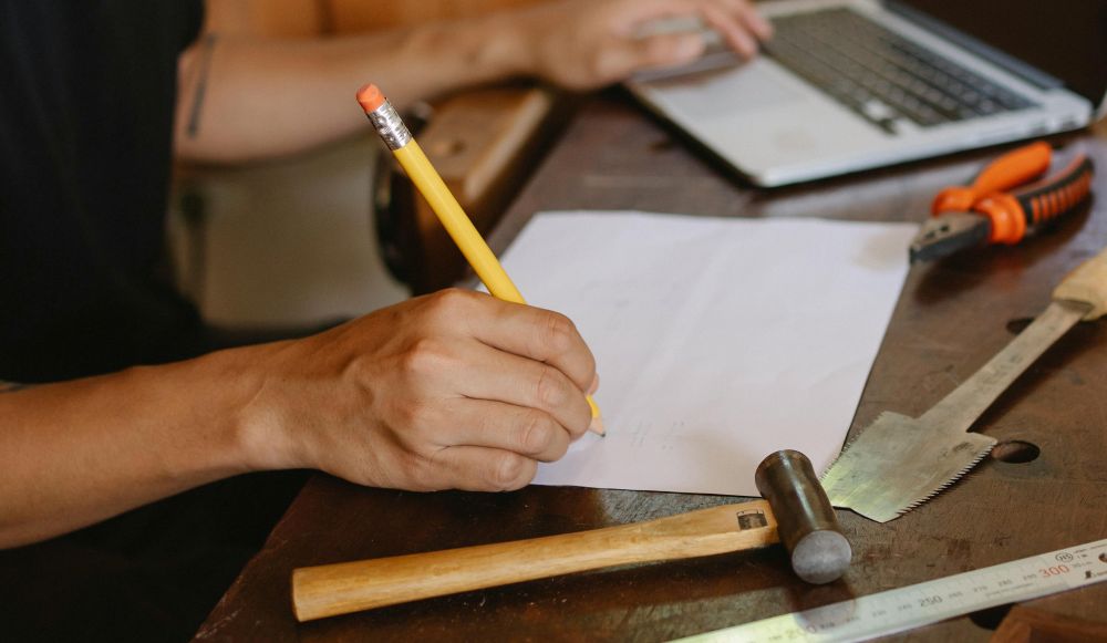 Handwerker skizziert auf Papier an einer Werkbank; Werkzeuge und Laptop liegen daneben.